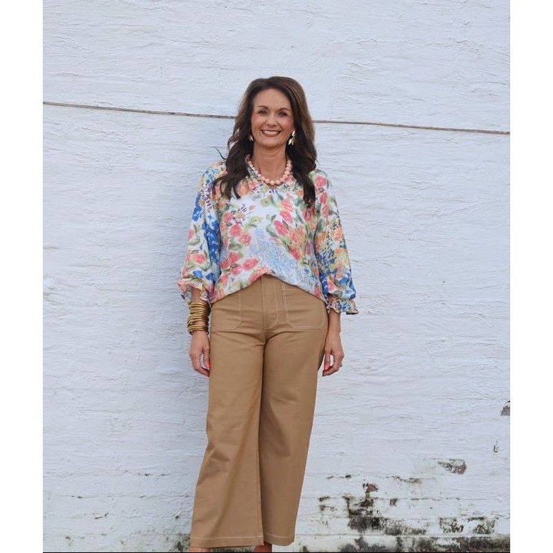 Woman standing against a white brick wall wearing a floral blouse and beige pants.