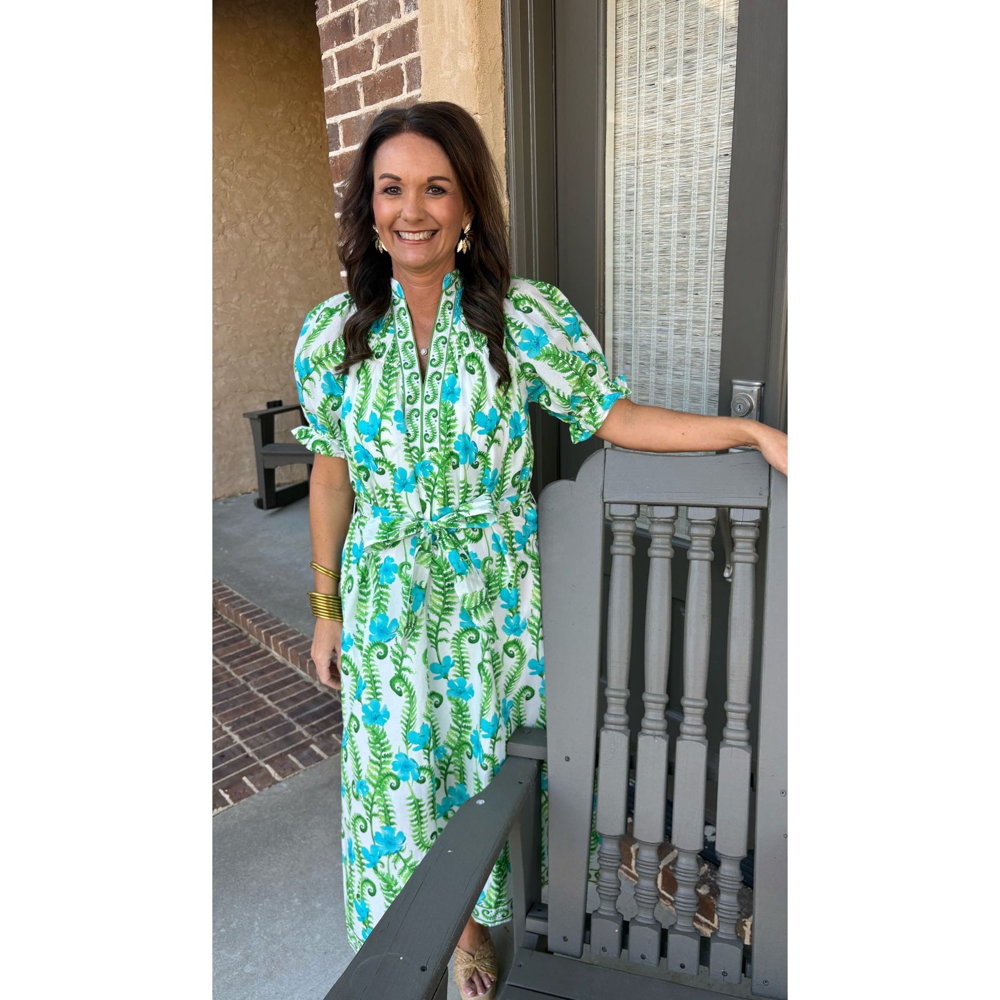 Woman in a green and blue floral dress standing on a porch.