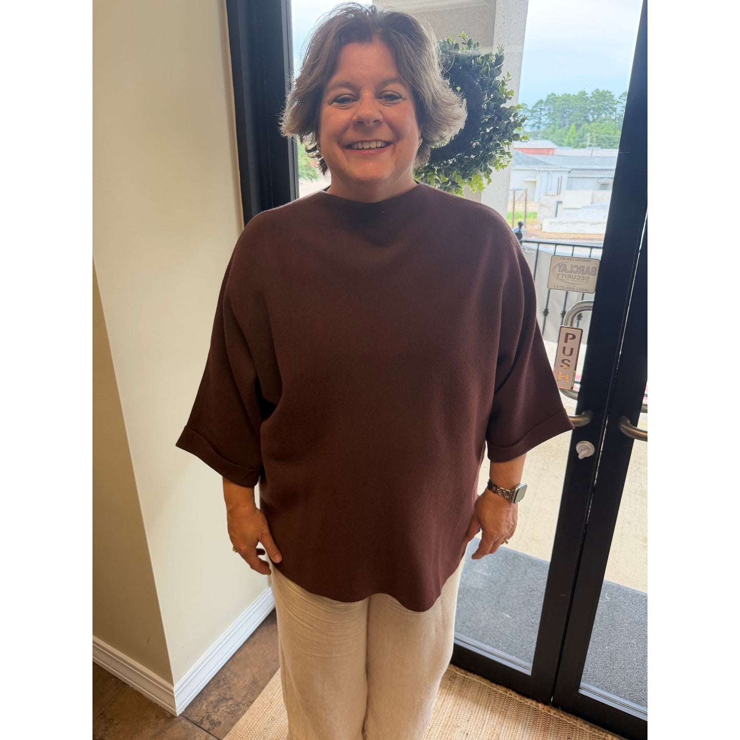 A smiling woman with short gray hair wearing an oversized dark brown mock-neck t-shirt and cream pants, standing indoors near a glass door.