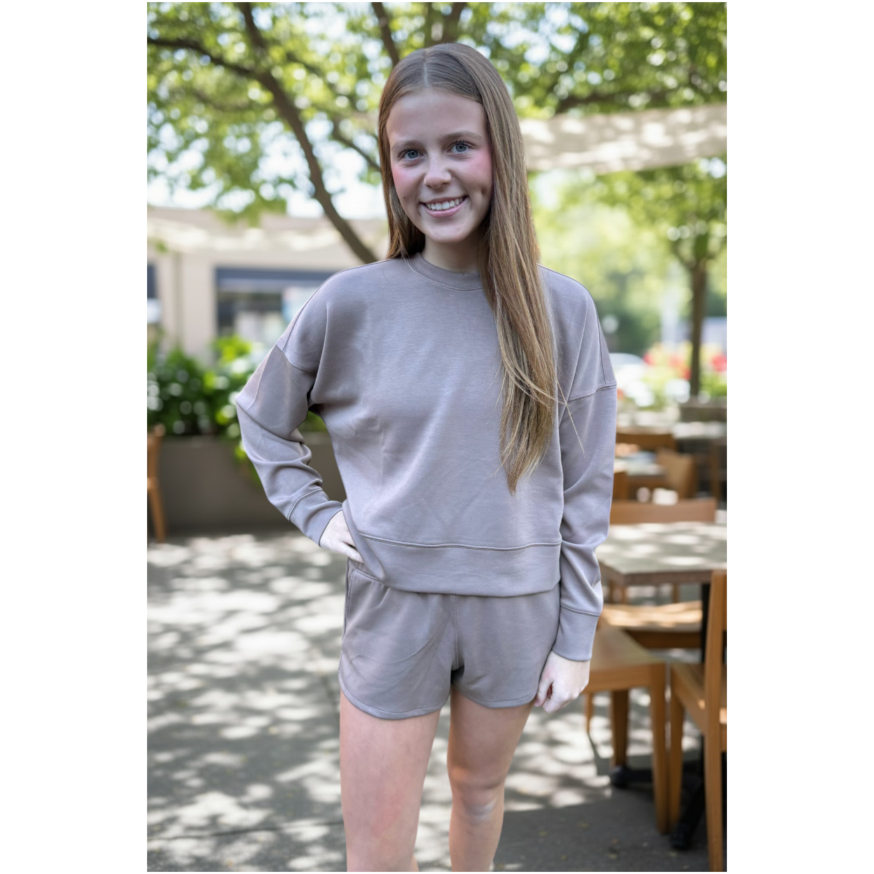 Person wearing a gray sweatshirt and shorts set outdoors with trees and tables in the background