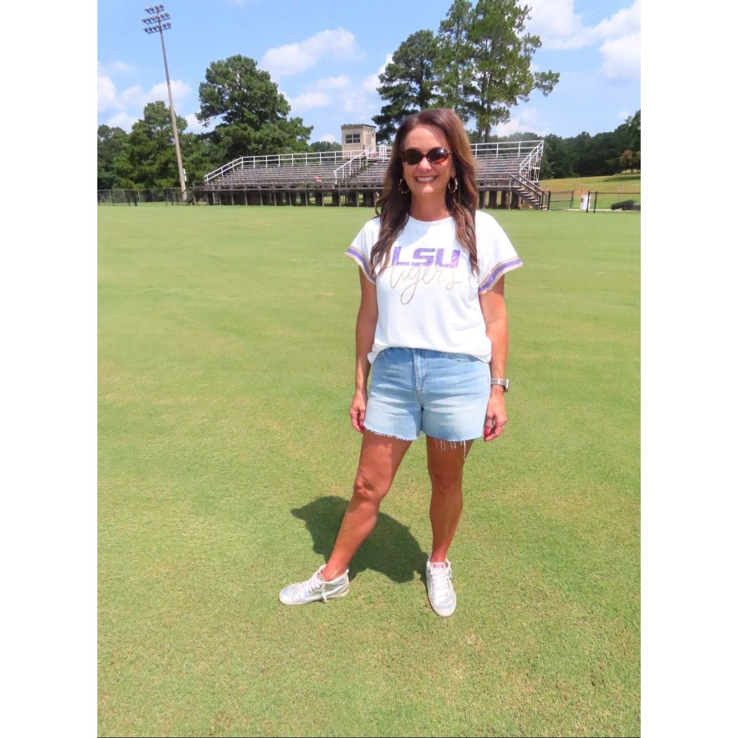 A woman stands on a grassy field wearing a white LSU Tigers graphic t-shirt, light blue denim shorts, and white sneakers, with stadium bleachers and trees in the background.