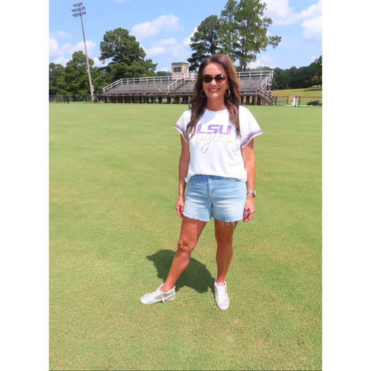A woman stands on a grassy field wearing a white LSU Tigers graphic t-shirt, light blue denim shorts, and white sneakers, with stadium bleachers and trees in the background.