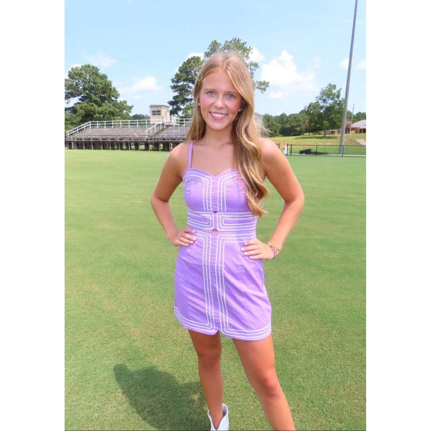 A woman stands on a green grass field wearing a lavender purple mini dress with white geometric embroidery. In the background, there is a grassy field, trees, and a building under a blue sky with clouds.