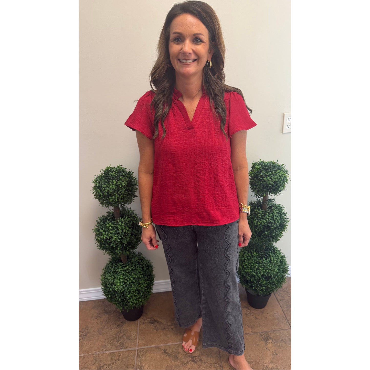 A woman stands indoors wearing a bright red short-sleeve textured top and dark gray wide-leg pants, with two small potted topiary trees on either side against a beige wall.