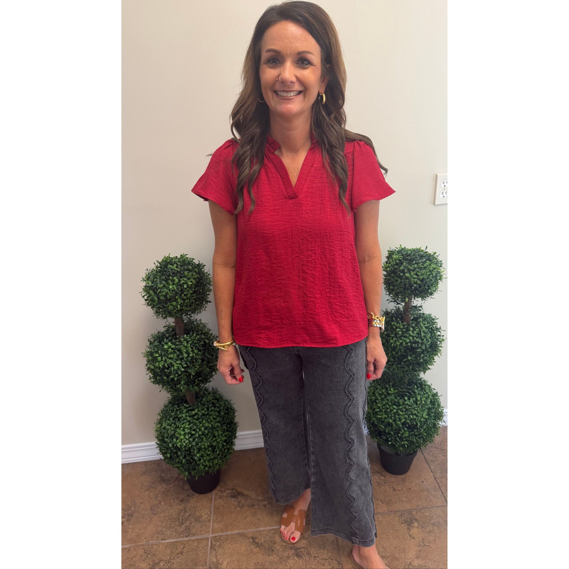 A woman stands indoors wearing a bright red short-sleeve textured top and dark gray wide-leg pants, with two small potted topiary trees on either side against a beige wall.