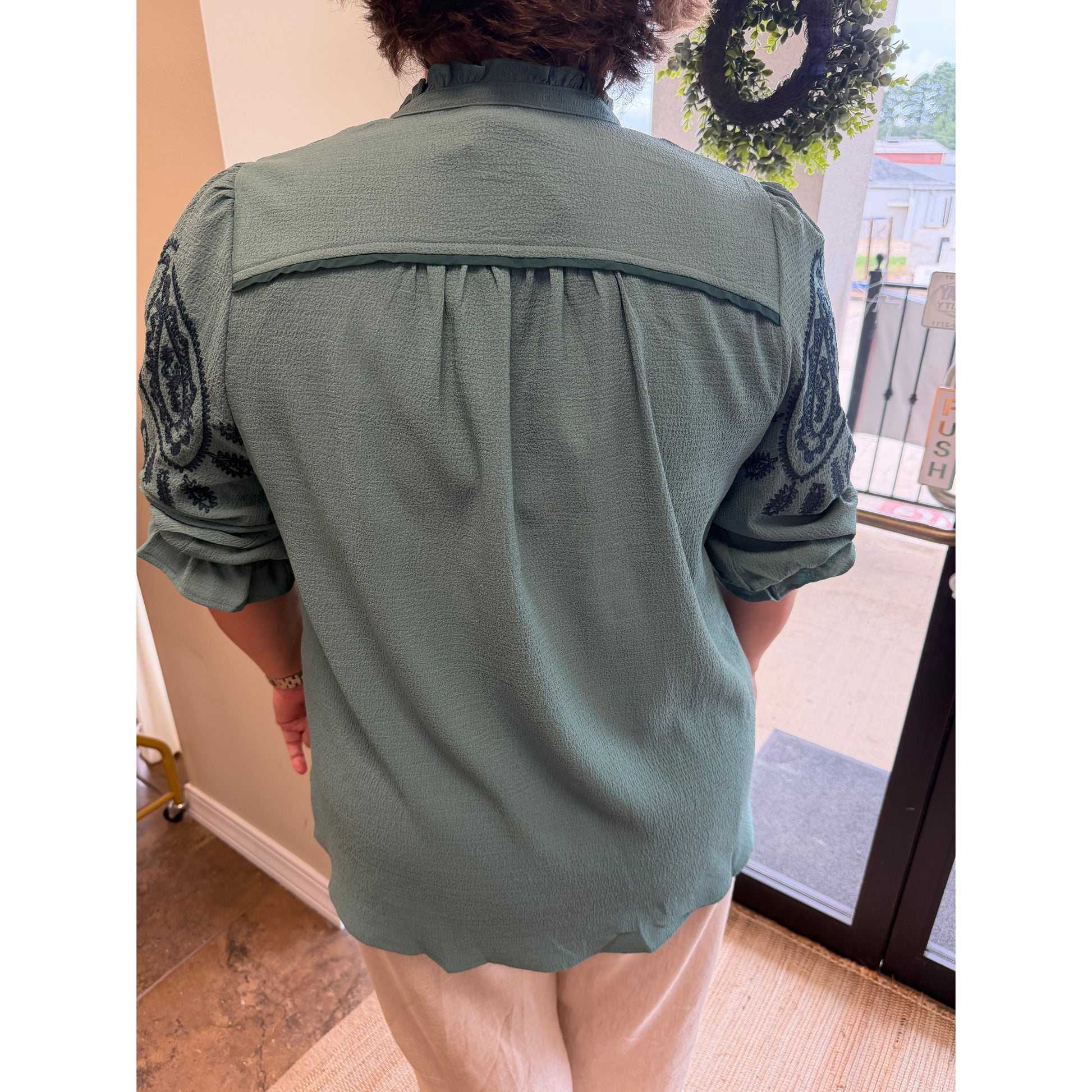 A person viewed from behind wearing a sage green blouse with dark embroidered detailing on the sleeves and a gathered yoke, paired with light beige pants in an indoor setting.