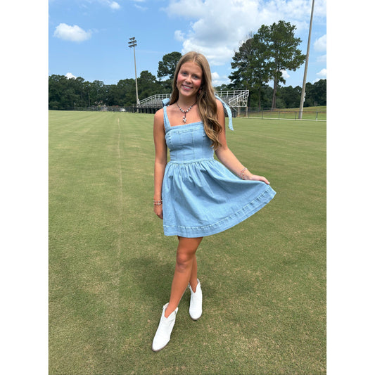 Woman in a blue dress standing on a grassy field with trees and a clear sky in the background.