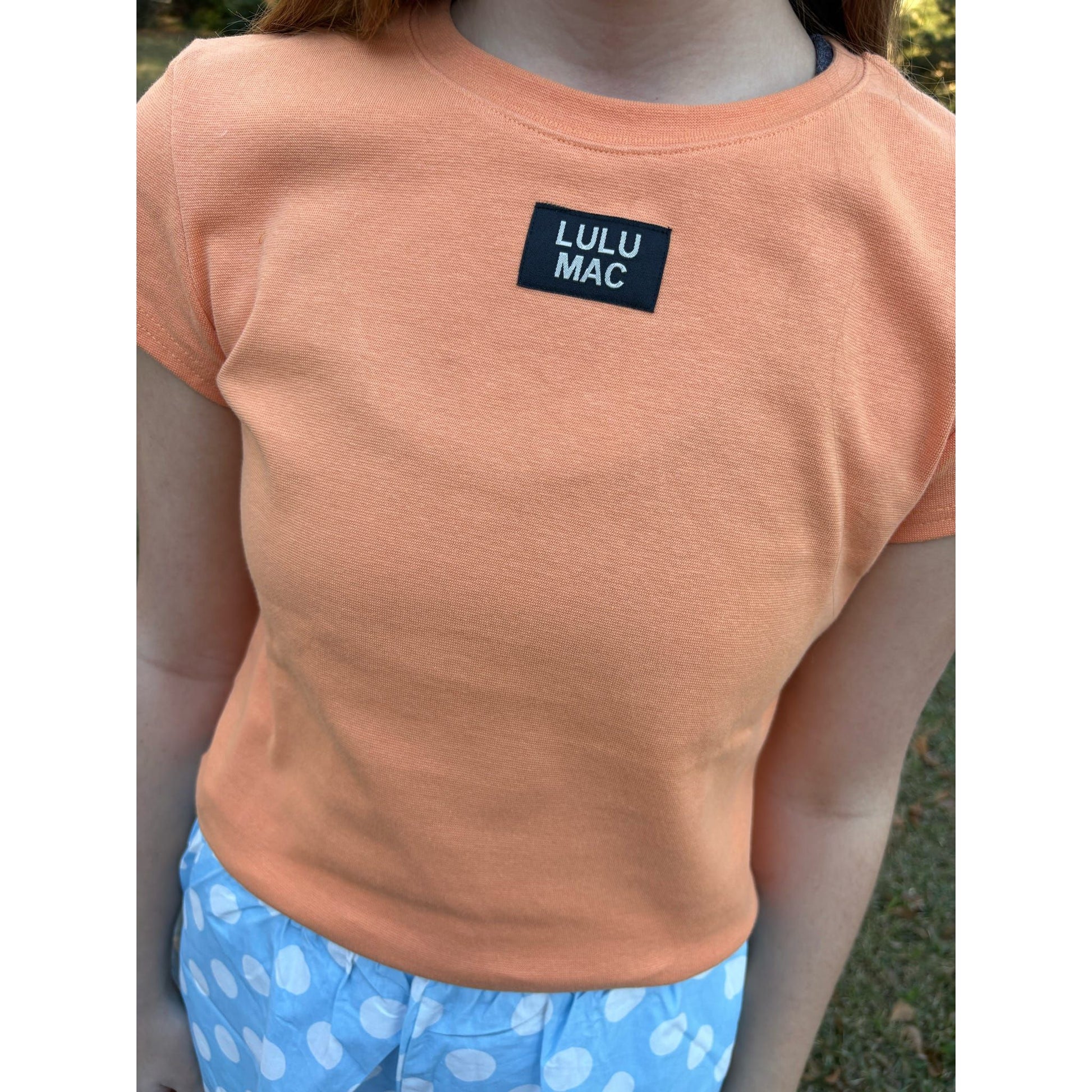 Person wearing an orange t-shirt with a 'LULU MAC' logo, standing outdoors.