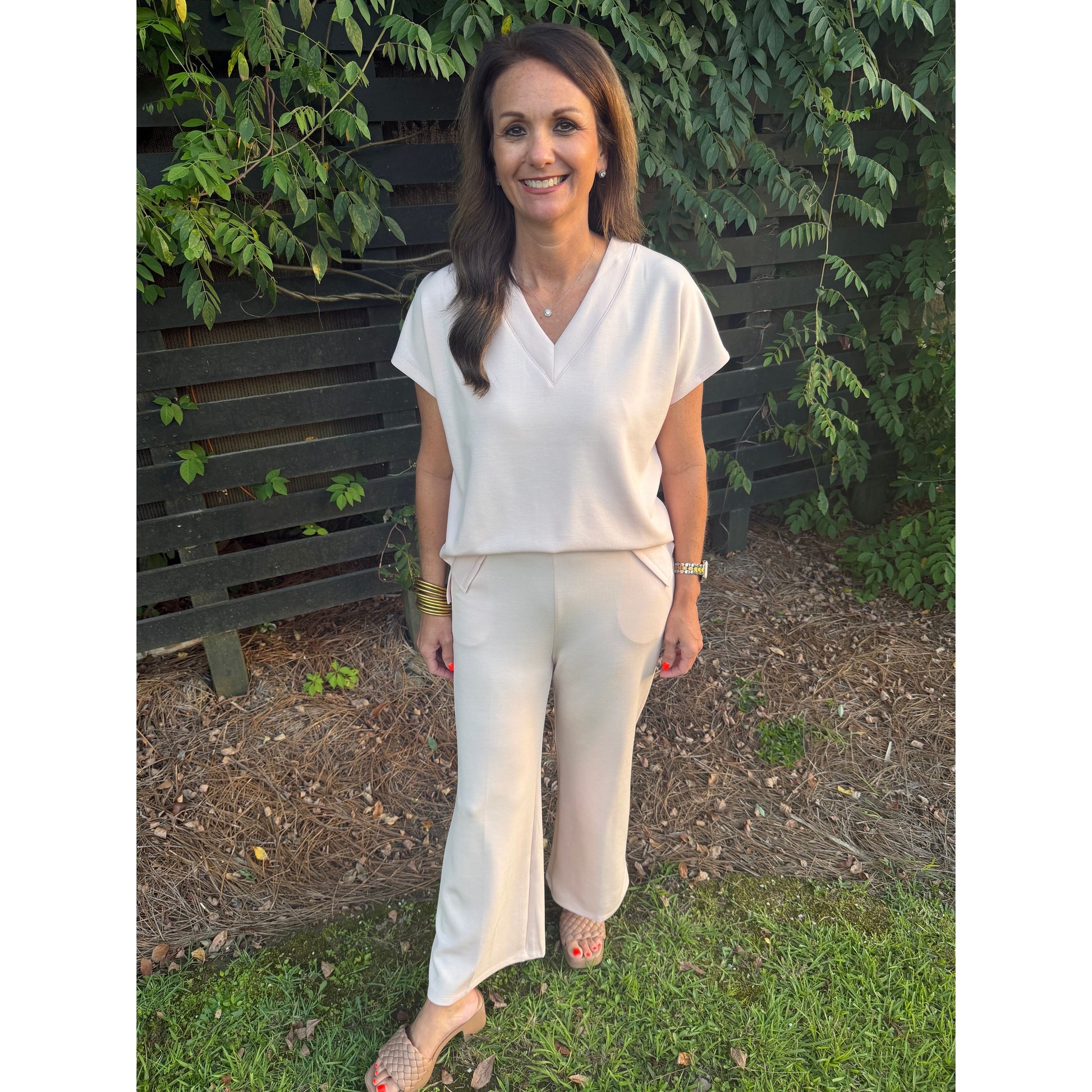 A woman stands outdoors in front of a dark wooden fence and greenery, wearing a light cream short-sleeve V-neck top and matching wide-leg pants with beige slingback sandals.