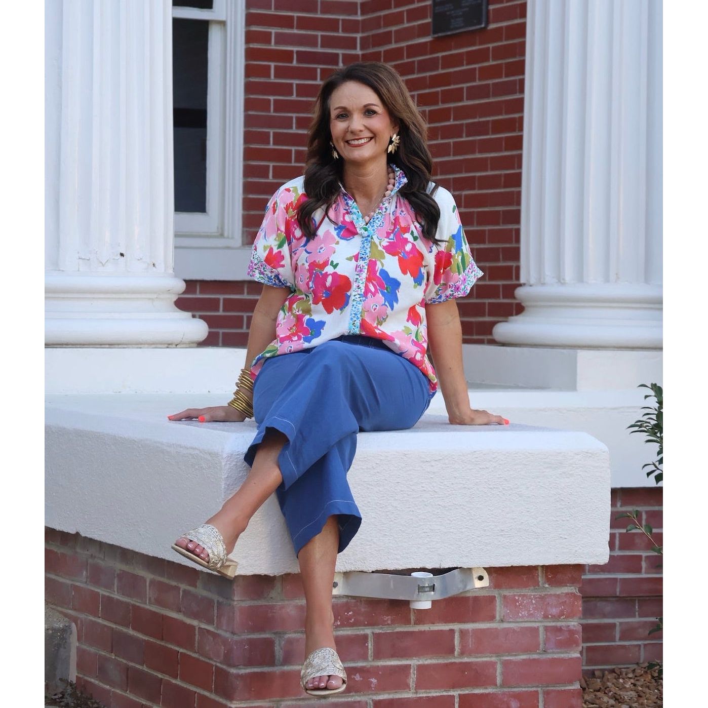 Woman sitting on a building ledge wearing a colorful floral blouse and blue pants.