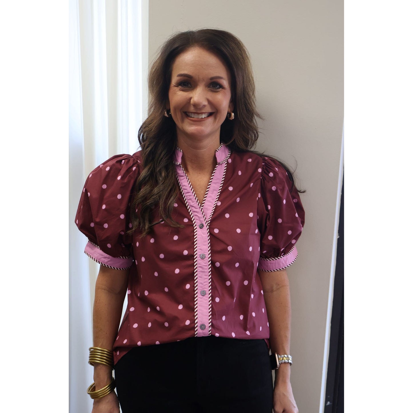 A smiling woman with long brown hair wearing a burgundy short-sleeve blouse with pink polka dots and pink trim, paired with black pants. She is standing indoors against a light-colored wall.