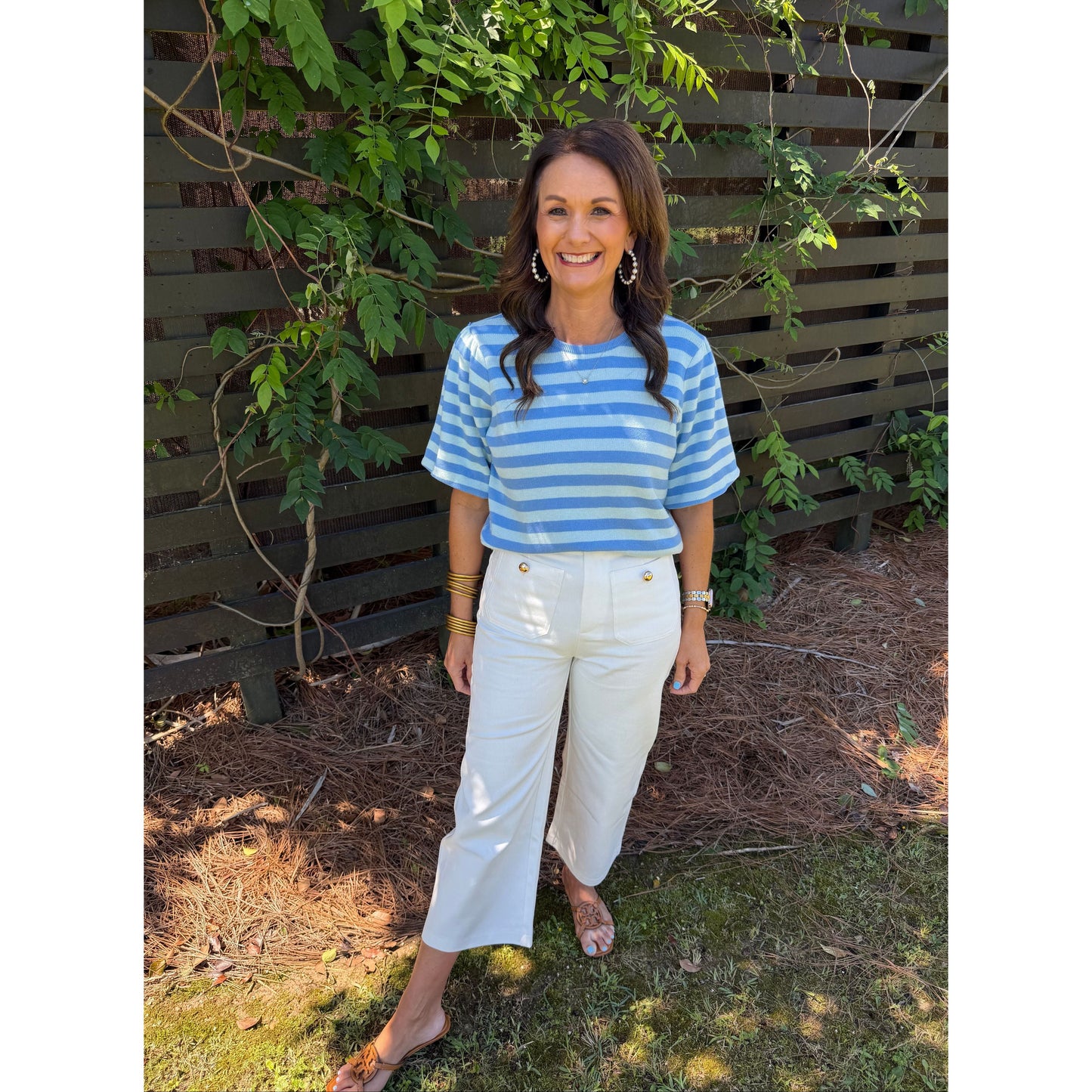 Woman in a blue and white striped shirt and white pants standing outdoors next to a wooden fence.