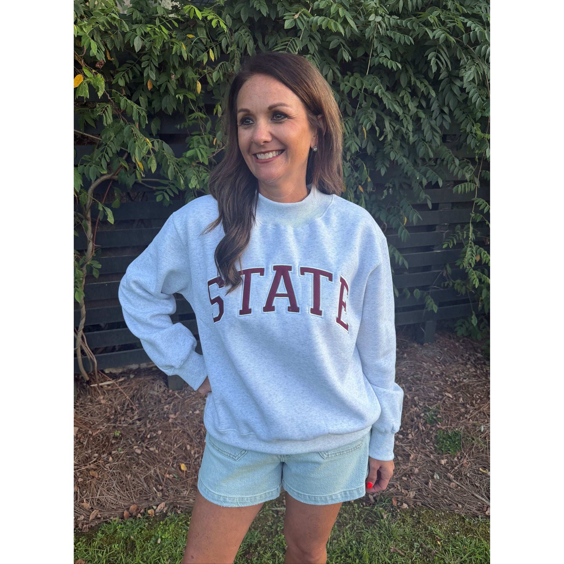 A woman wearing a light blue crewneck sweatshirt with '5 STATE' printed in maroon across the chest, paired with light denim shorts, standing outdoors in front of a green hedge.