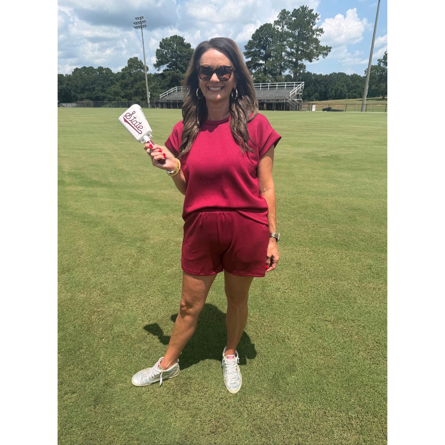 A woman wearing a matching burgundy short-sleeve top and shorts set stands on a green grass field, holding a white bottle with a red logo.