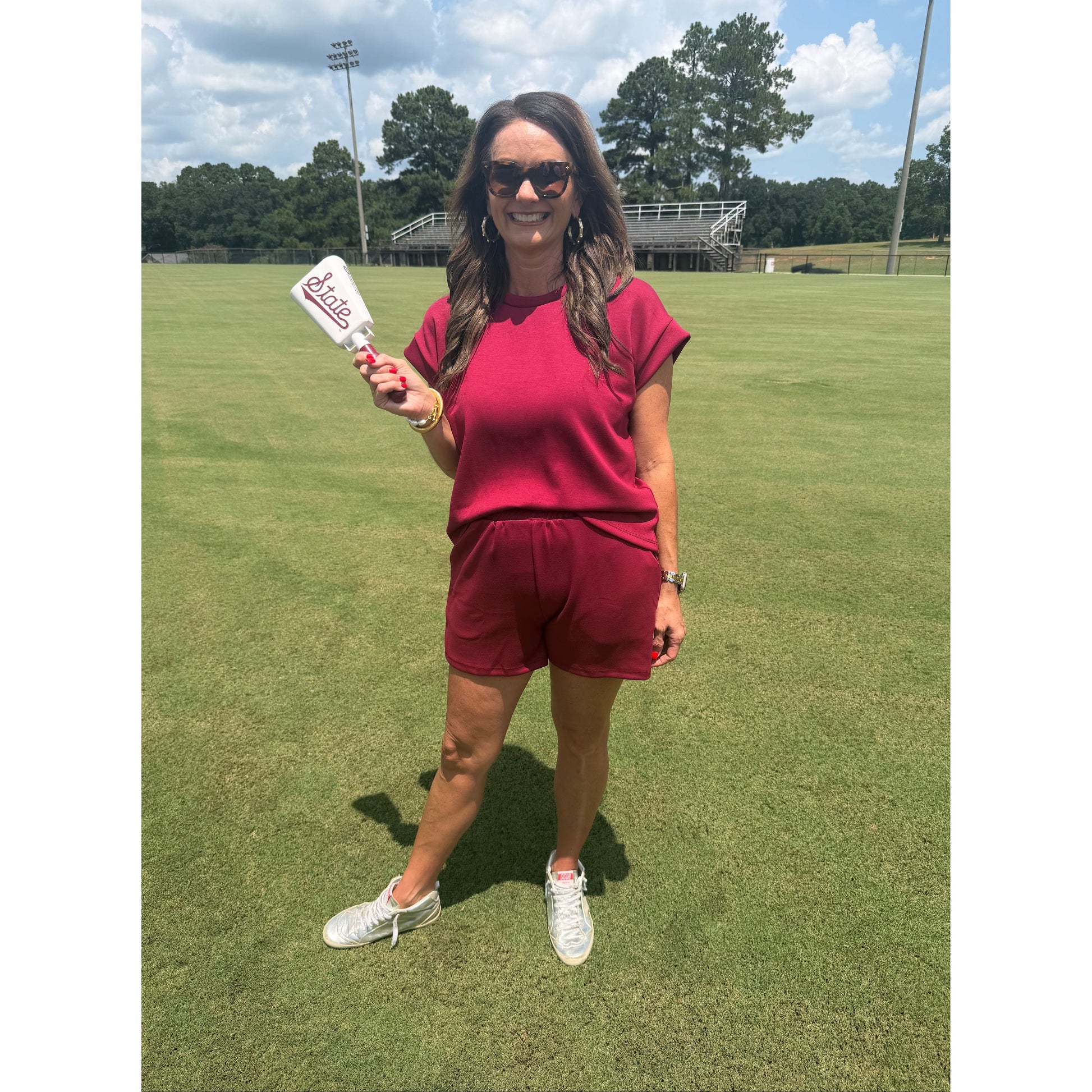A woman wearing a matching burgundy short-sleeve top and shorts set stands on a green grass field, holding a white bottle with a red logo.