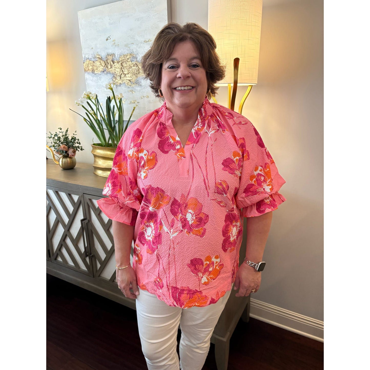 Woman wearing a pink floral blouse standing in a room with decor elements.