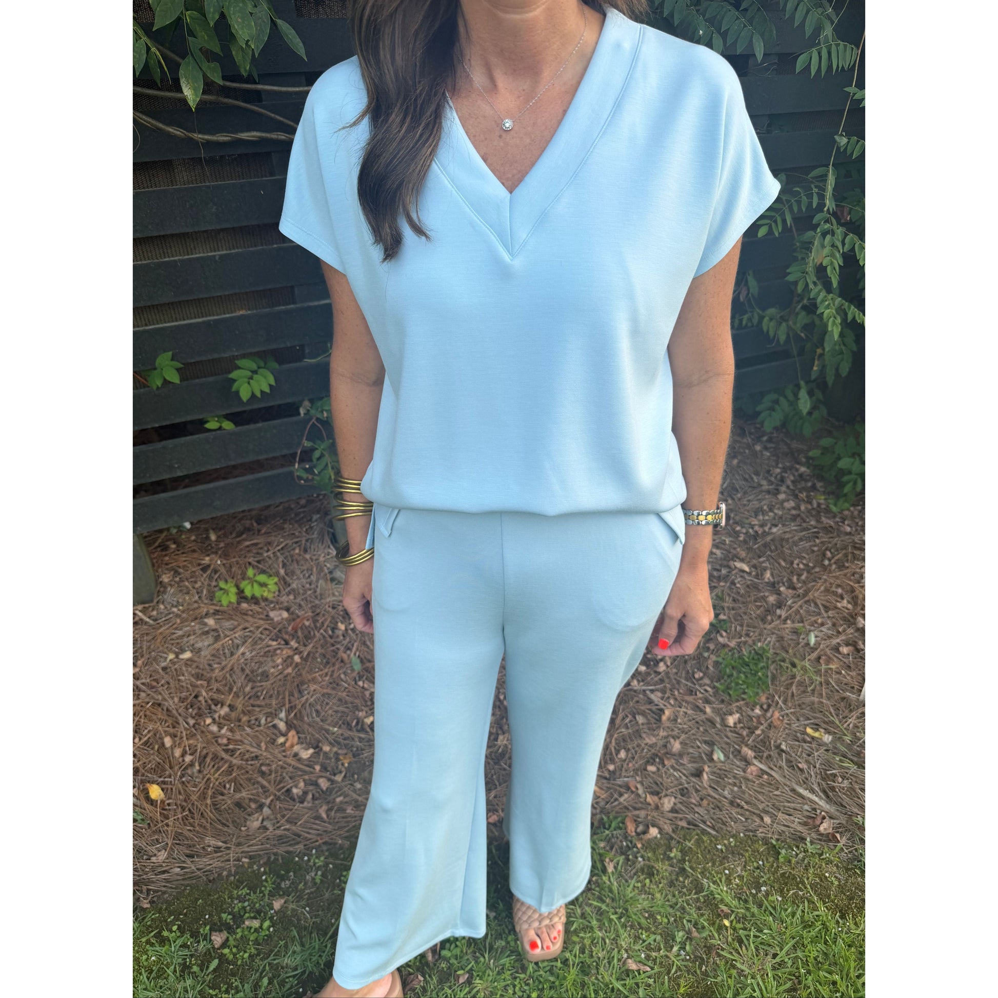 A woman wearing a light blue matching two-piece set consisting of a short-sleeve V-neck top and wide-leg pants, standing outdoors on grass in front of a dark wooden fence.