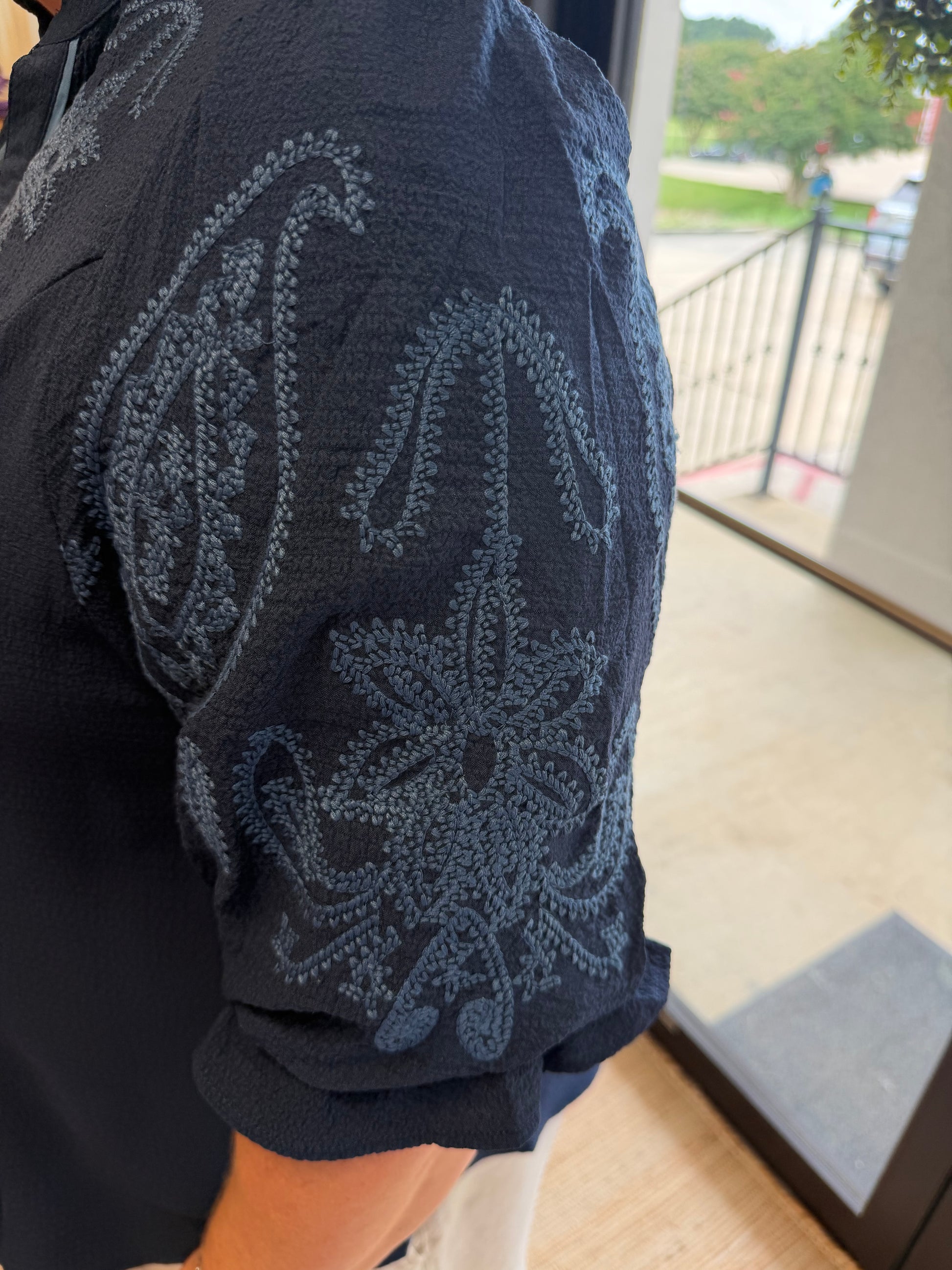 Close-up of a person wearing a dark navy short-sleeve shirt with a large tonal paisley pattern on the sleeve, standing indoors near a glass door.