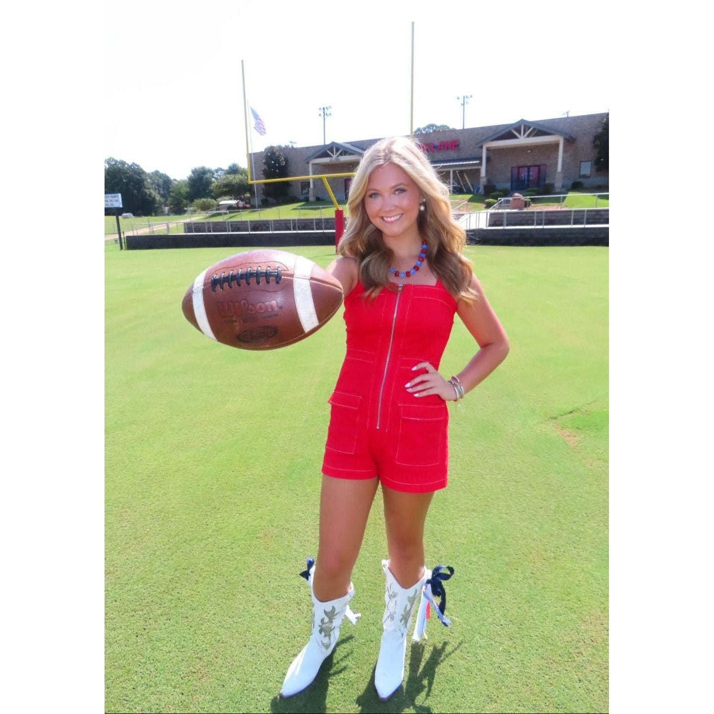 A woman stands on a green grass field holding a brown football, wearing a bright red sleeveless romper with a front zipper and white lace-up boots. A building and trees are visible in the background.