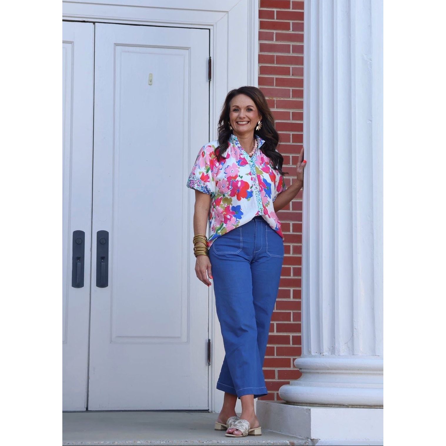 Woman in a colorful floral blouse and blue pants standing in front of a brick building with white doors.