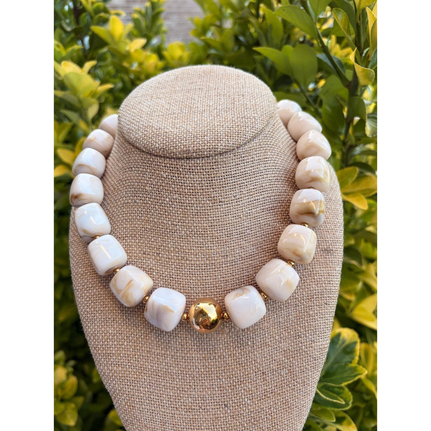 A chunky beaded necklace made of cream-colored stone beads with a central gold accent bead, displayed on a beige fabric jewelry bust against a background of green foliage.