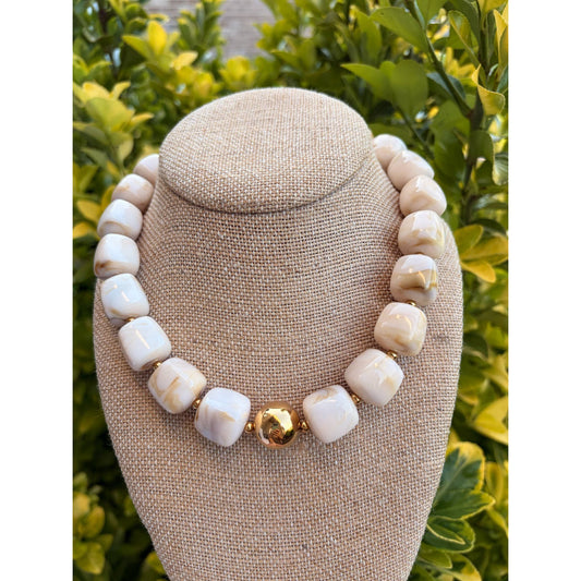 A chunky beaded necklace made of cream-colored stone beads with a central gold accent bead, displayed on a beige fabric jewelry bust against a background of green foliage.