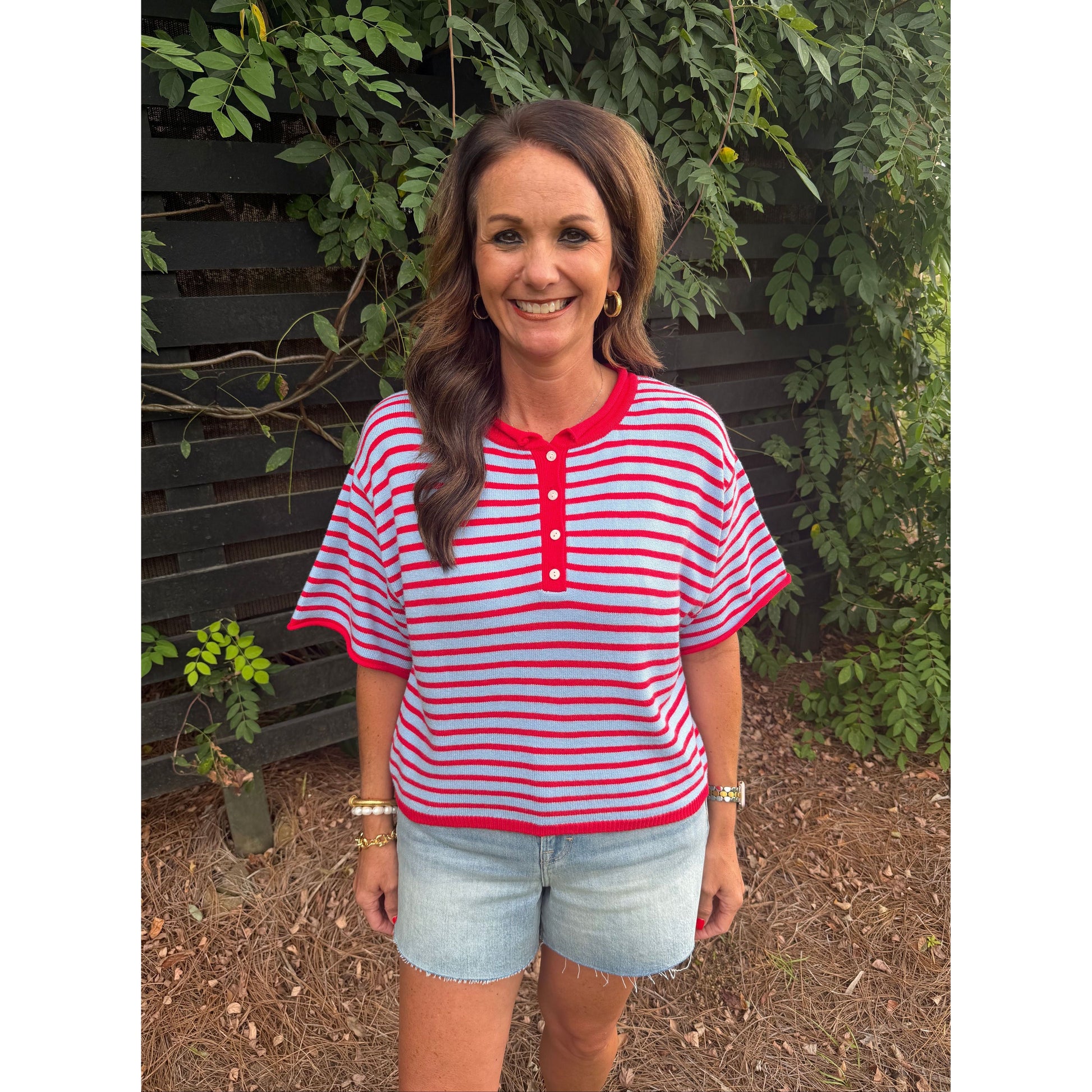 A woman wearing a red and white striped short-sleeve henley top with light blue denim shorts, standing outdoors in front of a dark wooden fence and green foliage.