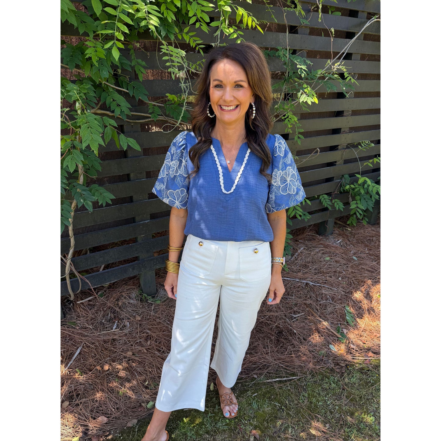 Woman wearing a blue blouse and white pants standing outdoors with greenery in the background