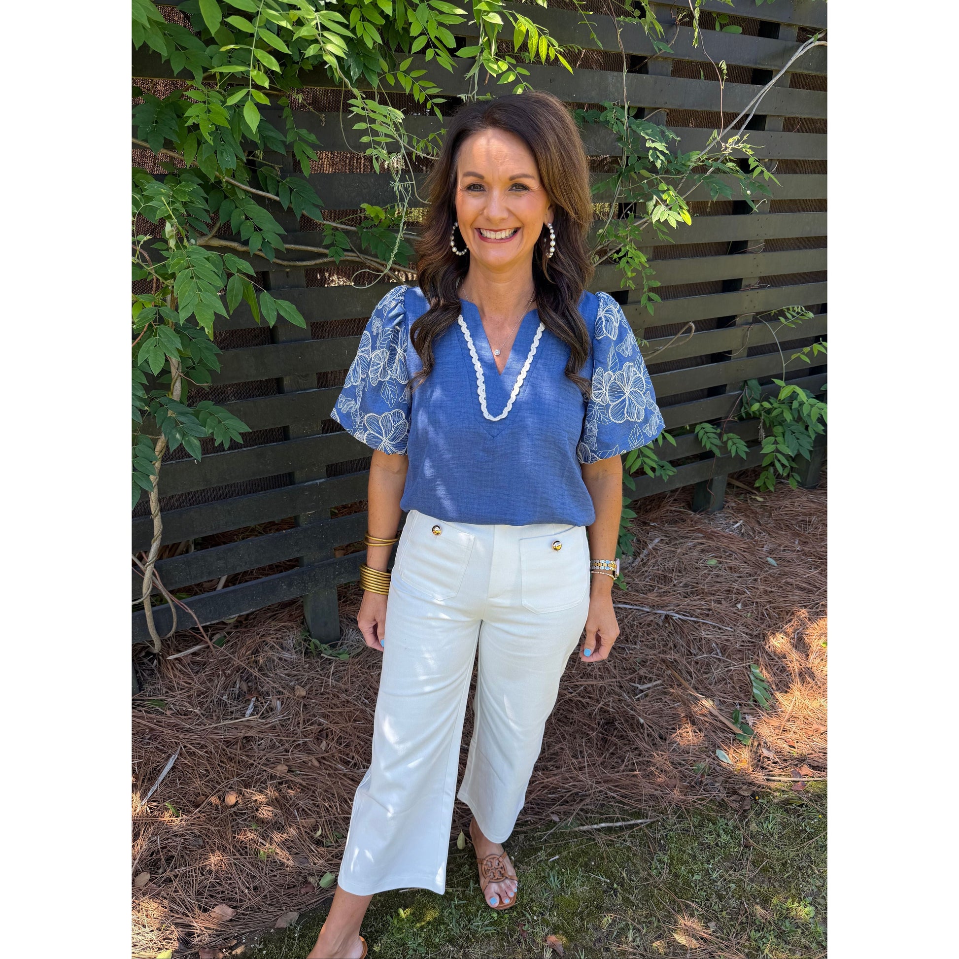 Woman wearing a blue blouse and white pants standing outdoors with greenery in the background