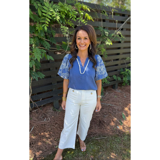 Woman wearing a blue blouse and white pants standing outdoors with greenery in the background