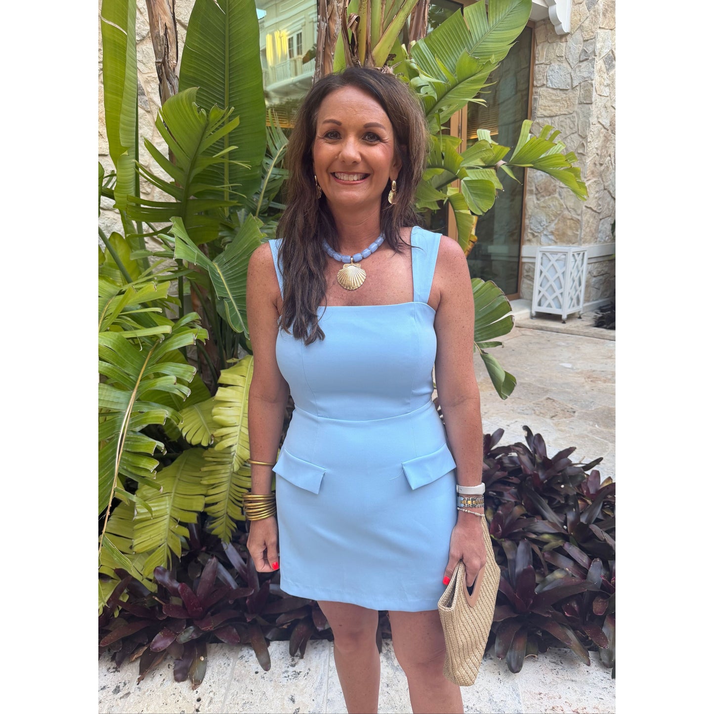 A woman wearing a light blue sleeveless mini dress with square shoulders and flap pockets, standing outdoors in front of tropical green plants and a stone wall.