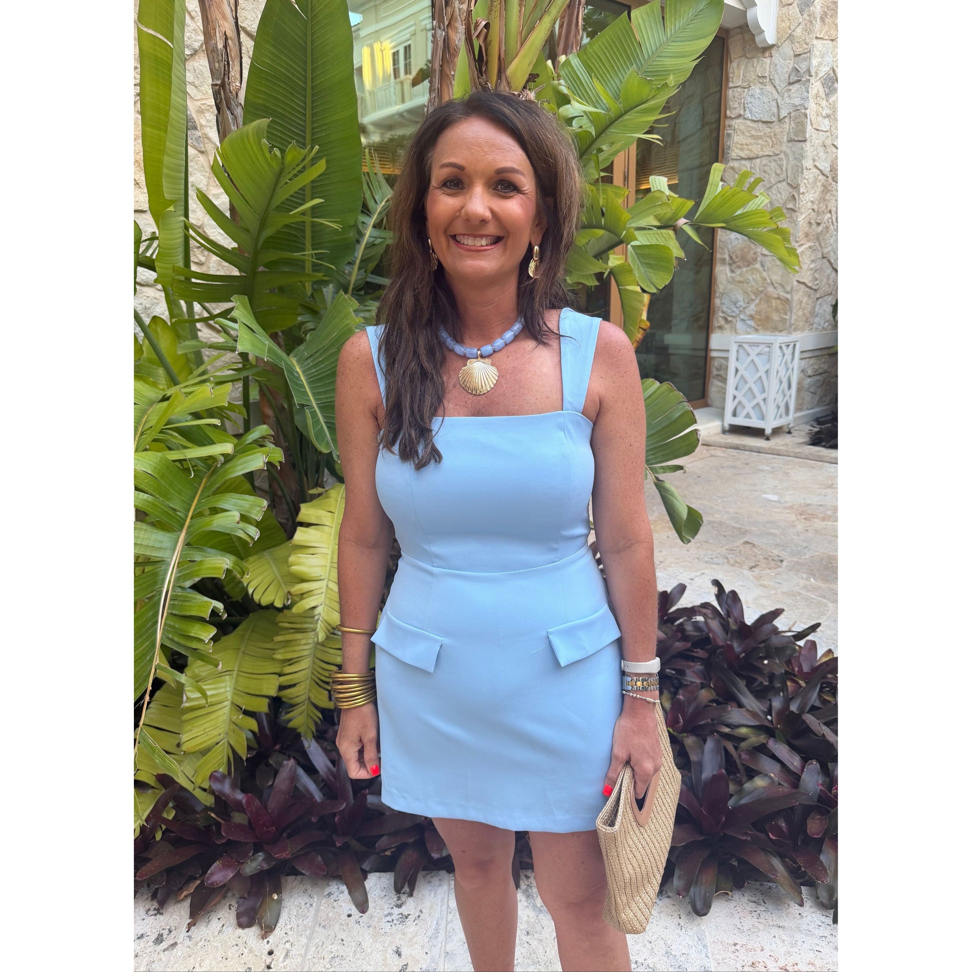 A woman wearing a light blue sleeveless mini dress with square shoulders and flap pockets, standing outdoors in front of tropical green plants and a stone wall.