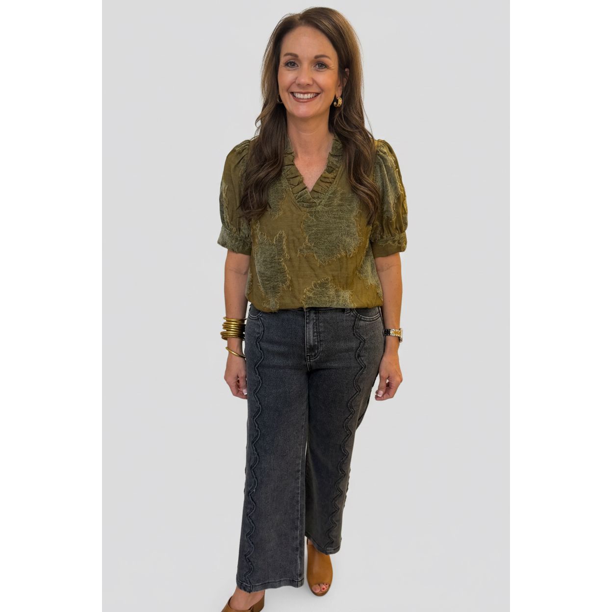 A woman stands indoors wearing a short-sleeve olive green patterned blouse and dark gray wide-leg jeans with snakeskin print details down the sides. She is wearing gold bracelets and tan flat shoes, standing in a home hallway with a white column, tiled floor, and a lamp in the background.