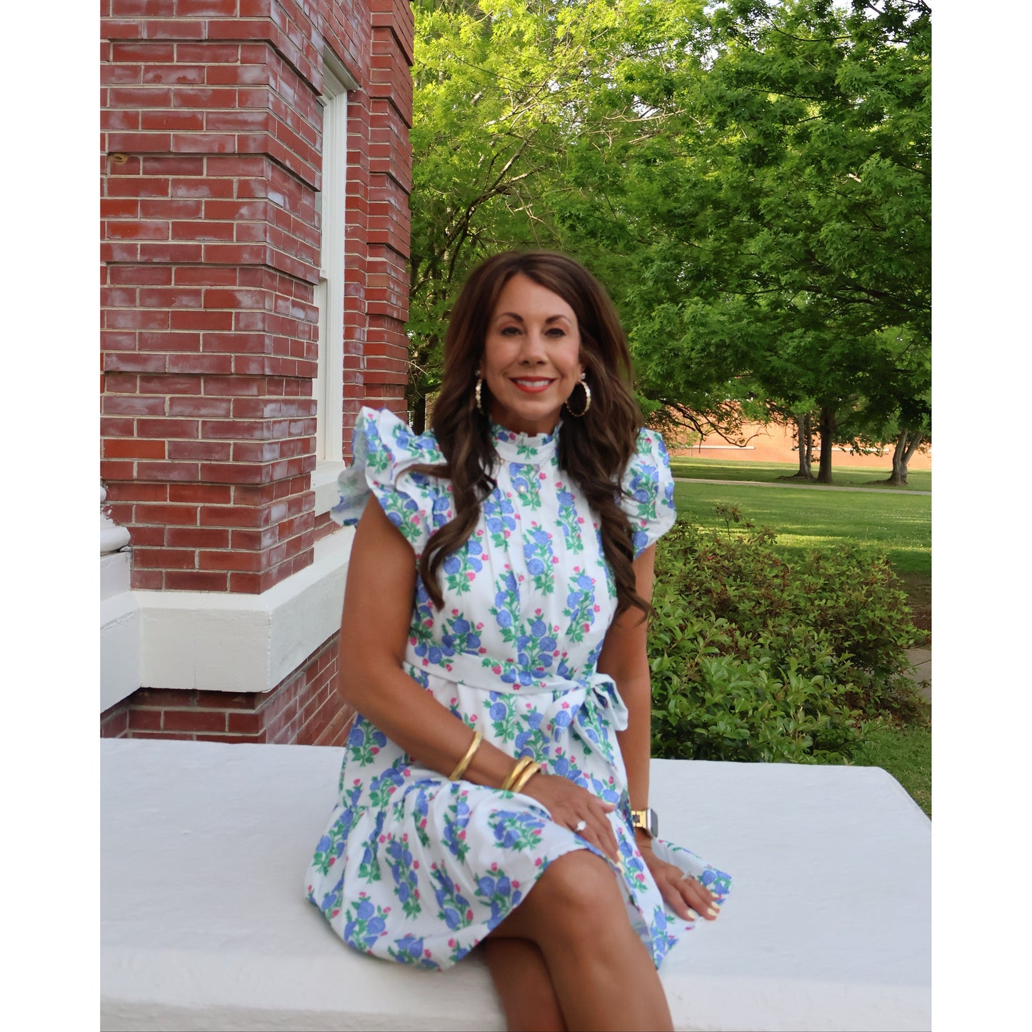 Young girl in a floral dress standing outdoors with trees and a brick wall in the background