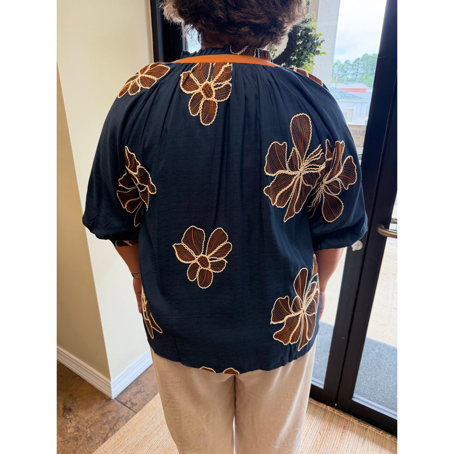 A person viewed from behind wearing a navy blue short-sleeve blouse with large brown and tan floral embroidery, paired with light beige pants, standing indoors near a glass door.