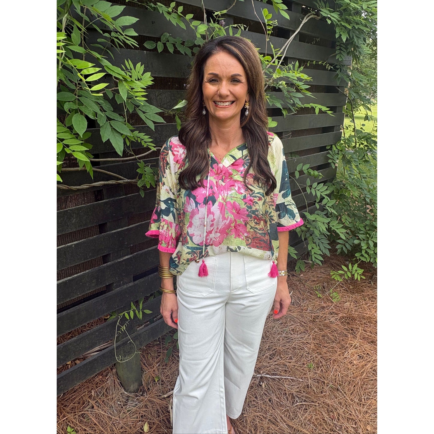 Woman wearing a floral blouse and white pants standing outdoors with greenery around.
