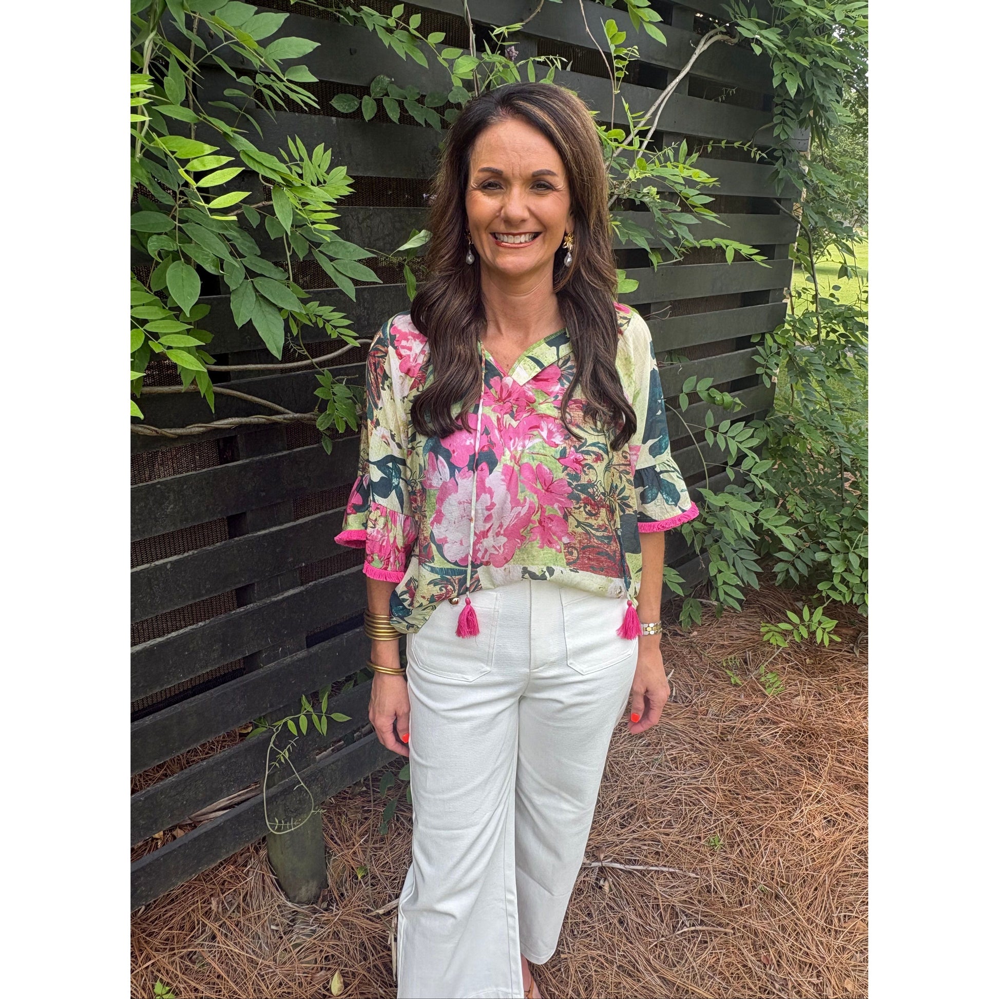Woman wearing a floral blouse and white pants standing outdoors with greenery around.
