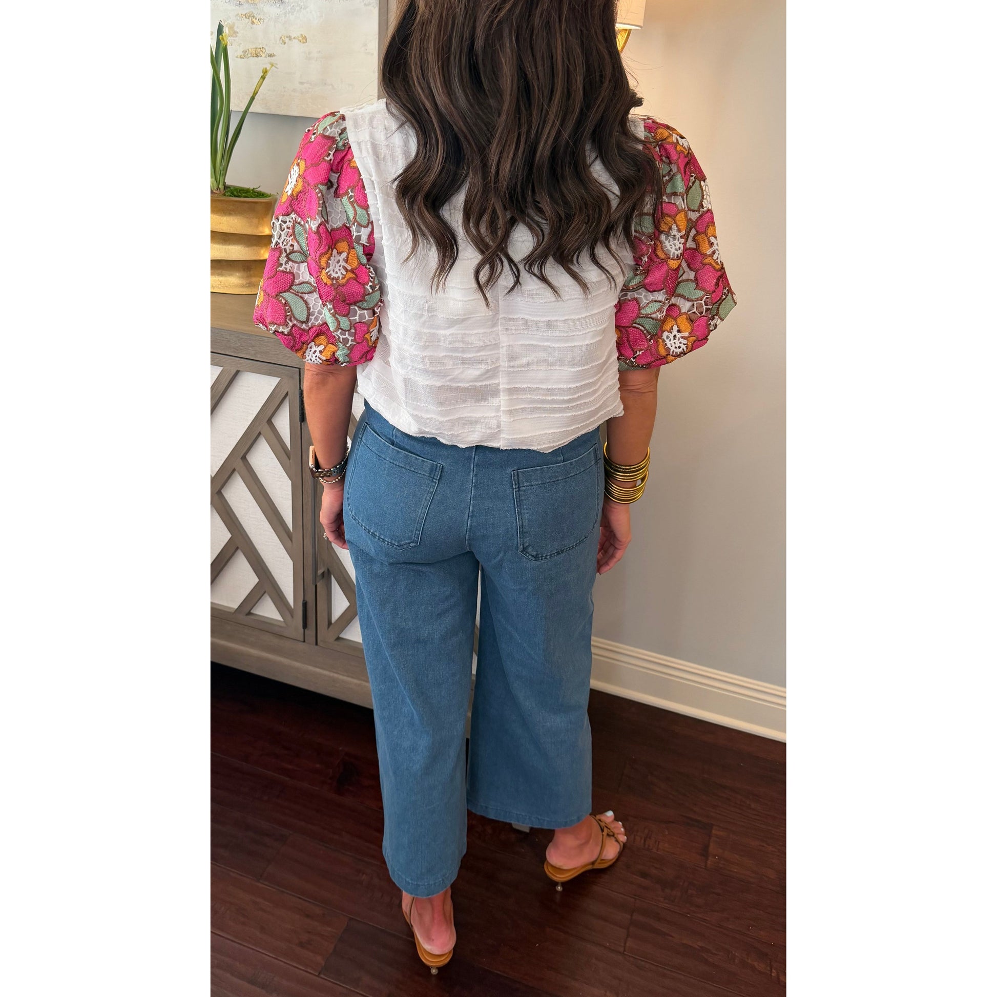 Person wearing a floral blouse and blue jeans standing in a room with a decorative cabinet and plant.