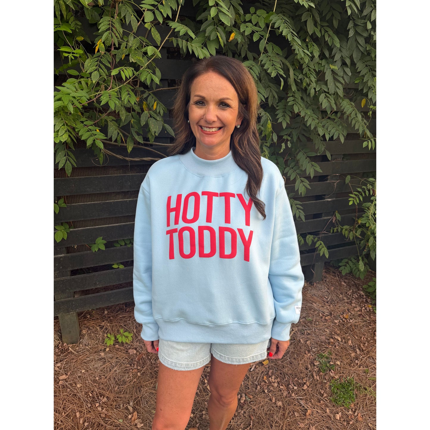 A woman wearing a light blue crewneck sweatshirt with 'HOTTY TODDY' printed in red across the chest, standing outdoors in front of a wooden fence and green foliage.