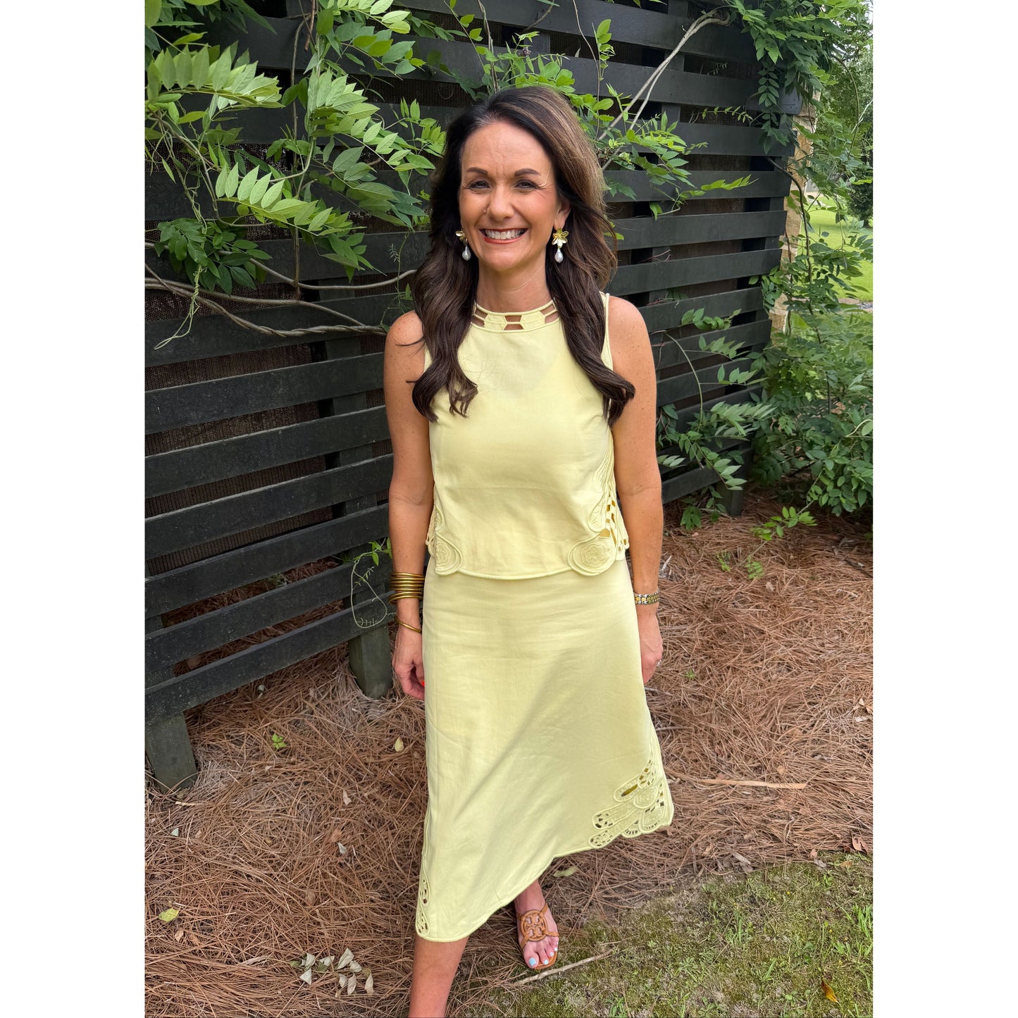 Woman in a yellow dress standing outdoors on a bench with greenery around.