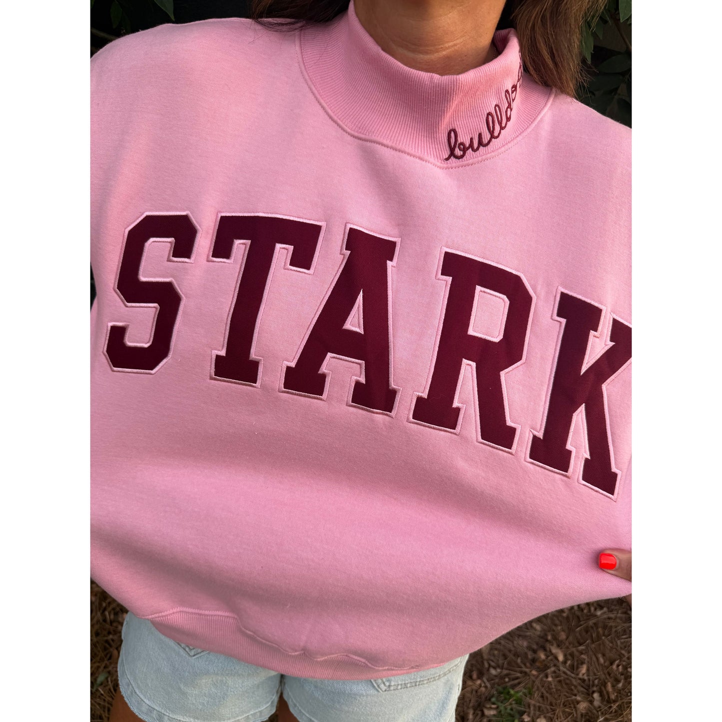 A close-up of a person wearing a light pink mock-neck sweatshirt with 'STARK' embroidered in dark maroon varsity letters across the chest. The brand name 'bulldog' is embroidered in black script on the inside of the collar.