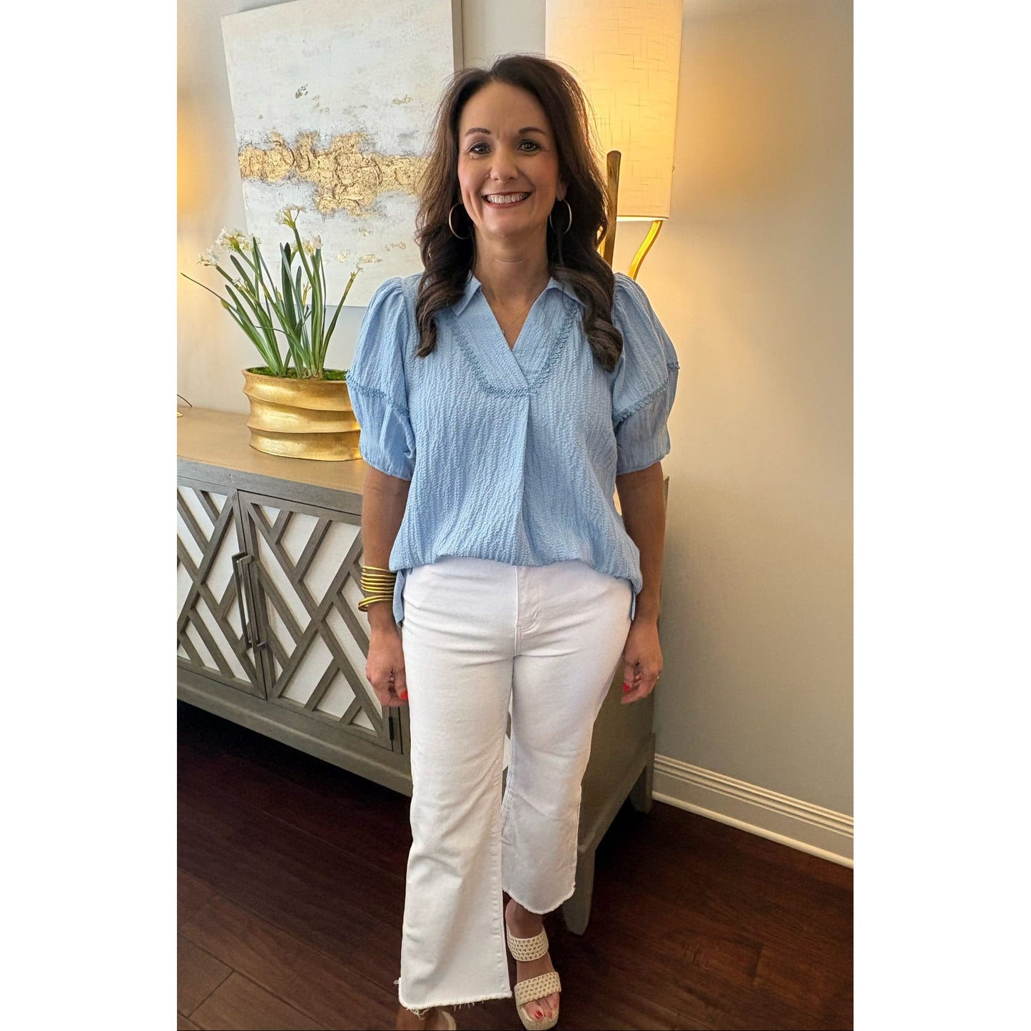 Woman in a blue top and white pants standing in a room with a lamp and decorative items.