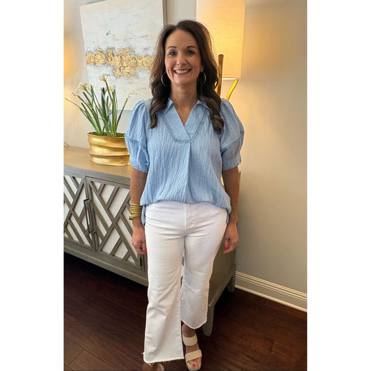Woman in a blue top and white pants standing in a room with a lamp and decorative items.