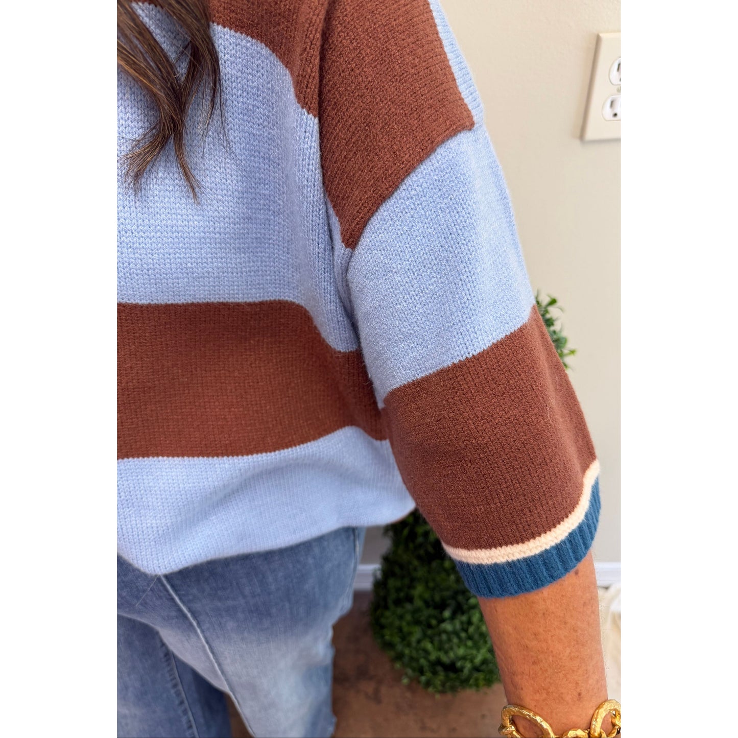 A close-up of a person wearing a knit sweater with wide horizontal stripes in light blue, rust brown, and cream, paired with blue jeans. The photo is cropped to show the torso and arm, with a beige wall, outlet, and small green plant in the background.