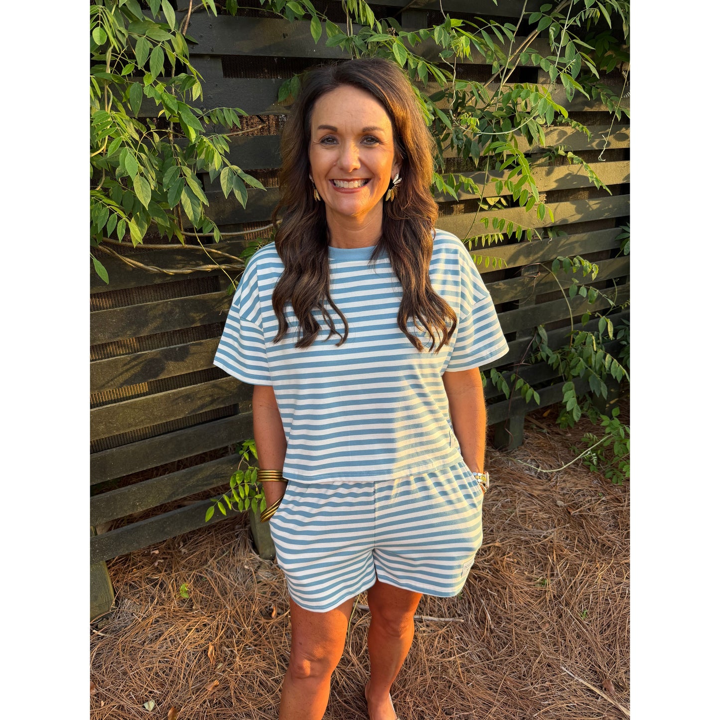 Woman wearing a blue and white striped dress standing in front of a wooden fence with greenery.