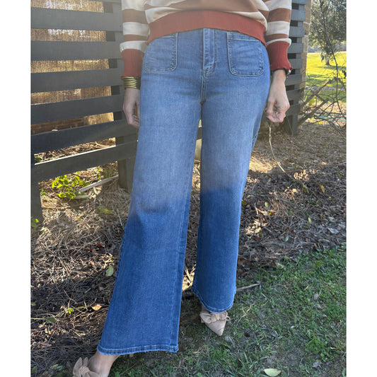 Person wearing a striped shirt and blue jeans sitting on a wooden bench outdoors.