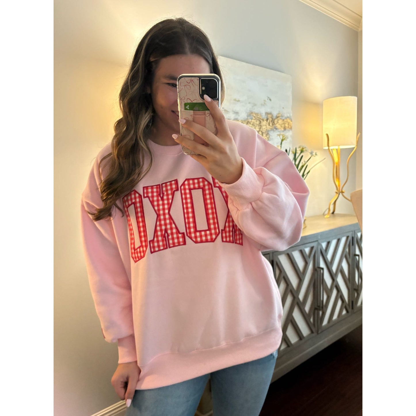 Person wearing a pink sweatshirt with red text, taking a mirror selfie in a room.
