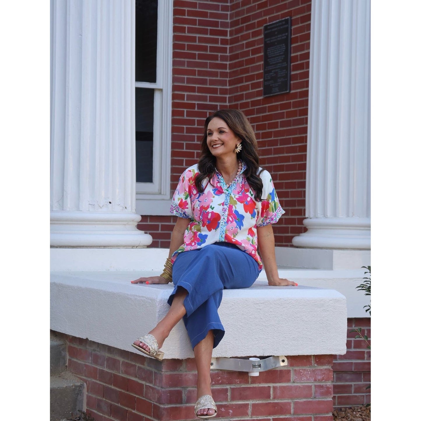 Woman sitting on a building's stone ledge wearing a colorful floral blouse and blue pants.