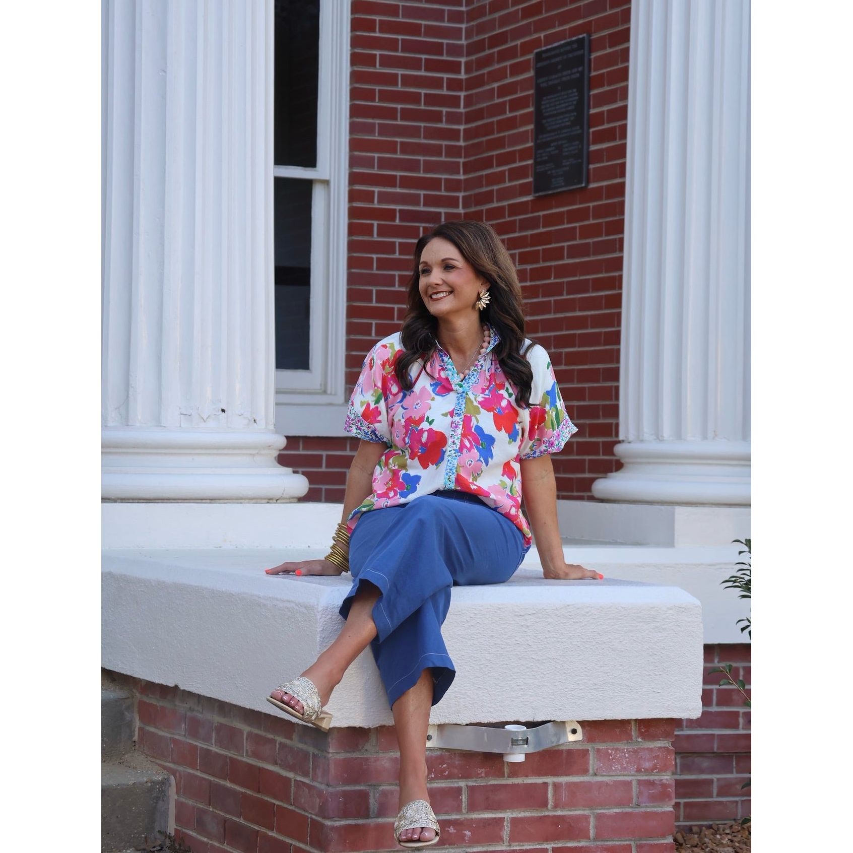 Woman sitting on a building's stone ledge wearing a colorful floral blouse and blue pants.