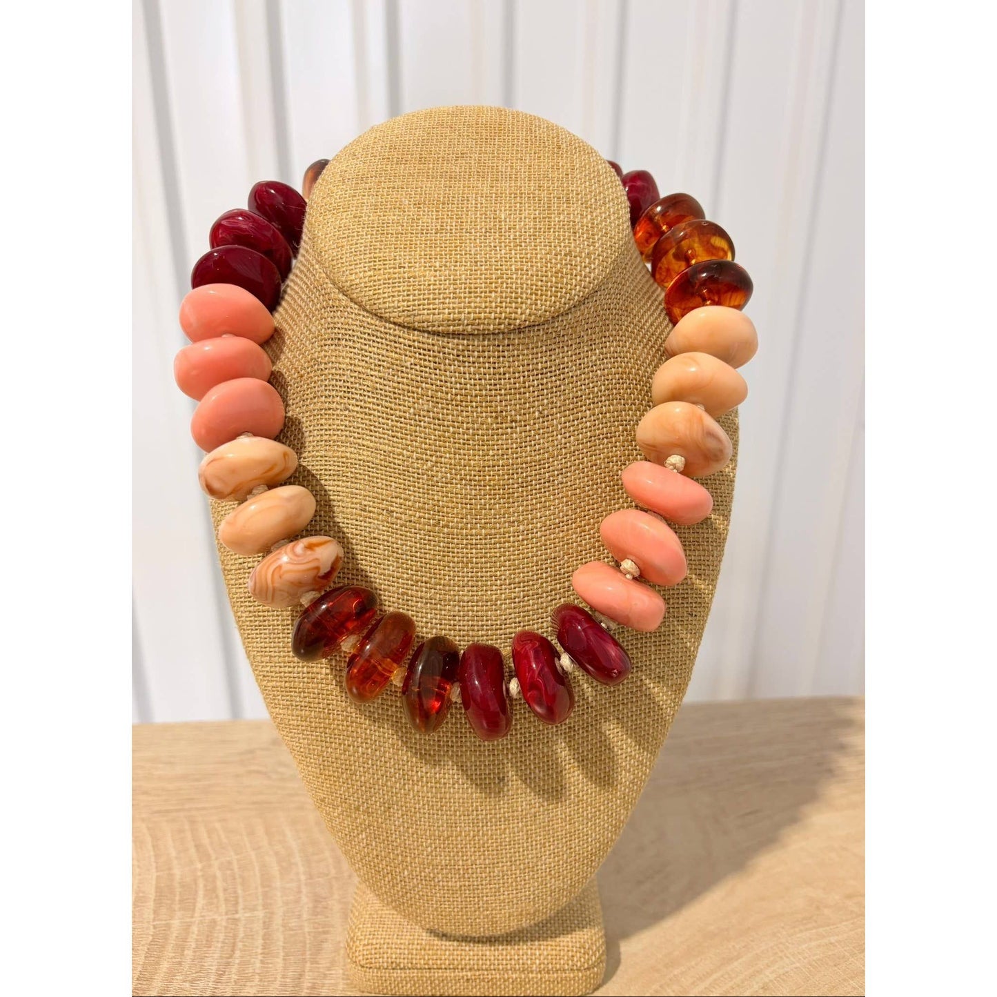 A chunky beaded necklace displayed on a tan fabric jewelry bust. The necklace features alternating strands of large polished coral-pink beads, deep red marbled beads, and translucent amber-orange beads, separated by tiny white spacer beads.